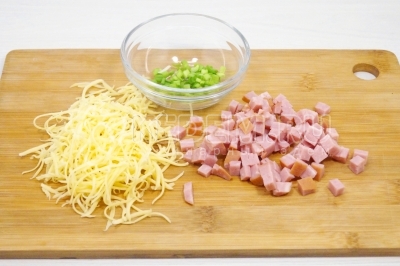 Чтобы приготовить омлет-конверт с ветчиной и сыром, нужно мелким кубиком нарезать 30 грамм ветчины. Натереть на мелкой терке 20 грамм сыра. Зеленый лук нашинковать.